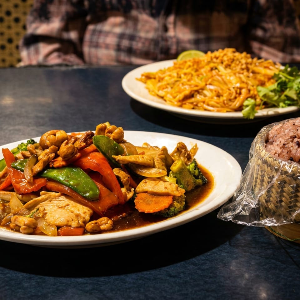 Plates of stir-fry, Pad Thai, and sticky rice in a bamboo container on a dark table.