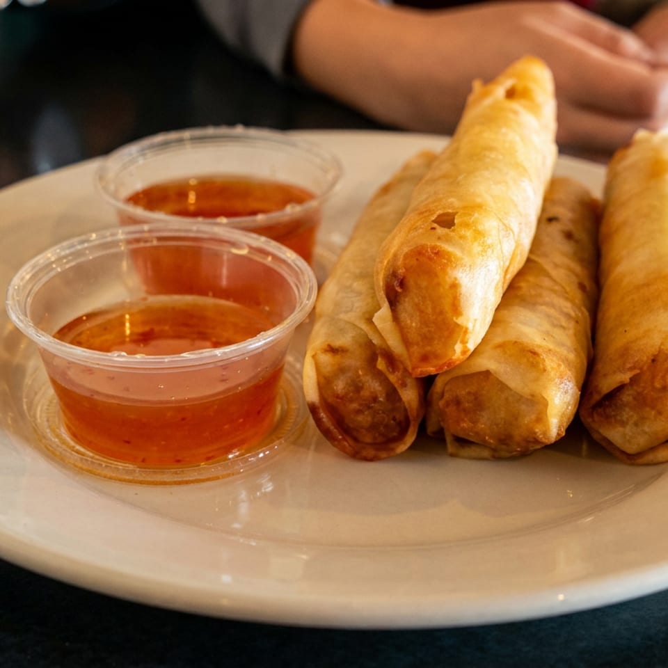 Four golden-brown spring rolls and two small cups of dipping sauce on a white plate.