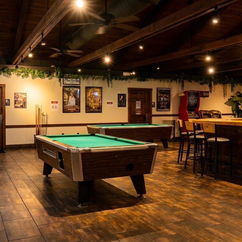 A dimly lit bar area with two pool tables, a wooden bar, and high-top seating.