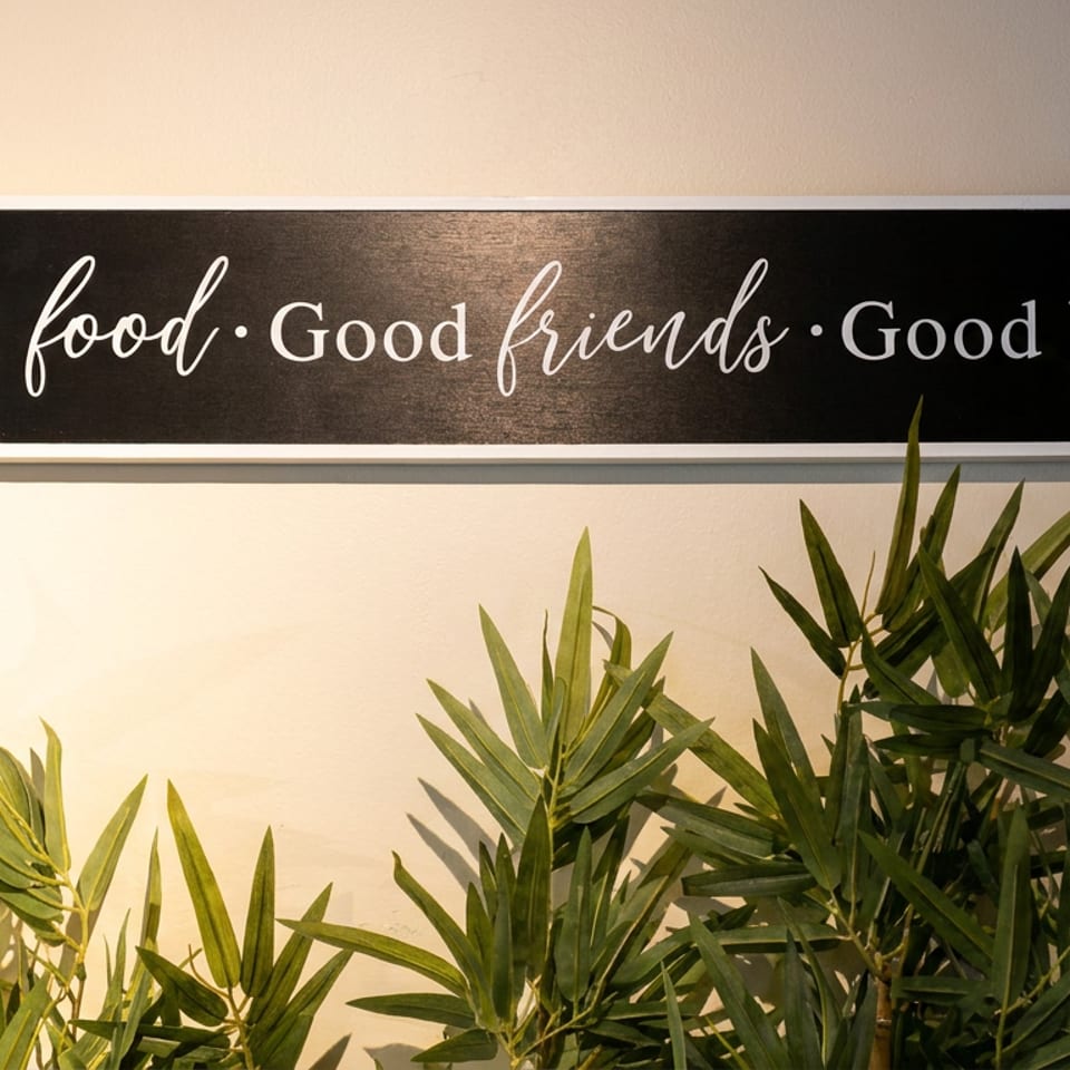 A black sign with white text "Good food • Good friends • Good times" hangs above green plants.